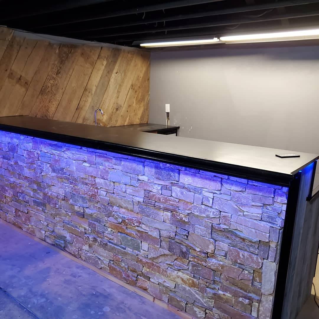 A finished basement bar with a dry-stacked stone front lit from below in blue, a dark counter top and a reclaimed timber wall behind it.