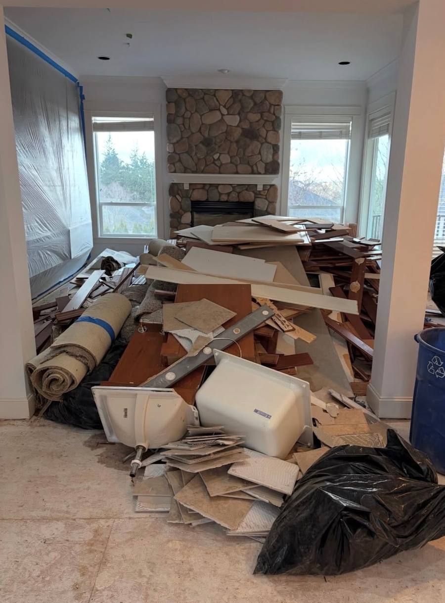 A room stripped out mid-remodel, with torn-up flooring, old cabinet carcasses and rolled carpet heaped in the middle of the floor in front of a stone fireplace and a bay window.