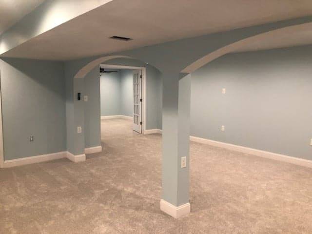 A finished basement room with pale grey walls, new carpet and a squared-off arched opening leading through to another room.